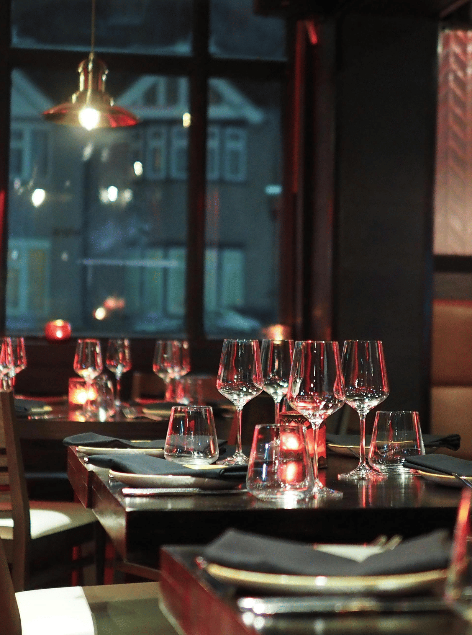 A moody interior shot of a restaurant dining table set with wine glasses and candles, seen through a dark window.