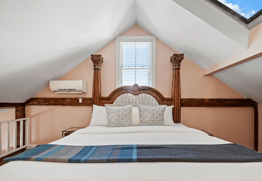 A bed with a wooden frame and white bedding is positioned under a skylight in an attic room with peach walls.