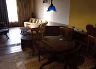 Living and dining area at The Bristol Hotel, Gurgaon with a round wooden dining table, 4 carved chairs, a cream sofa, a blue pendant lamp, and tiled flooring.
