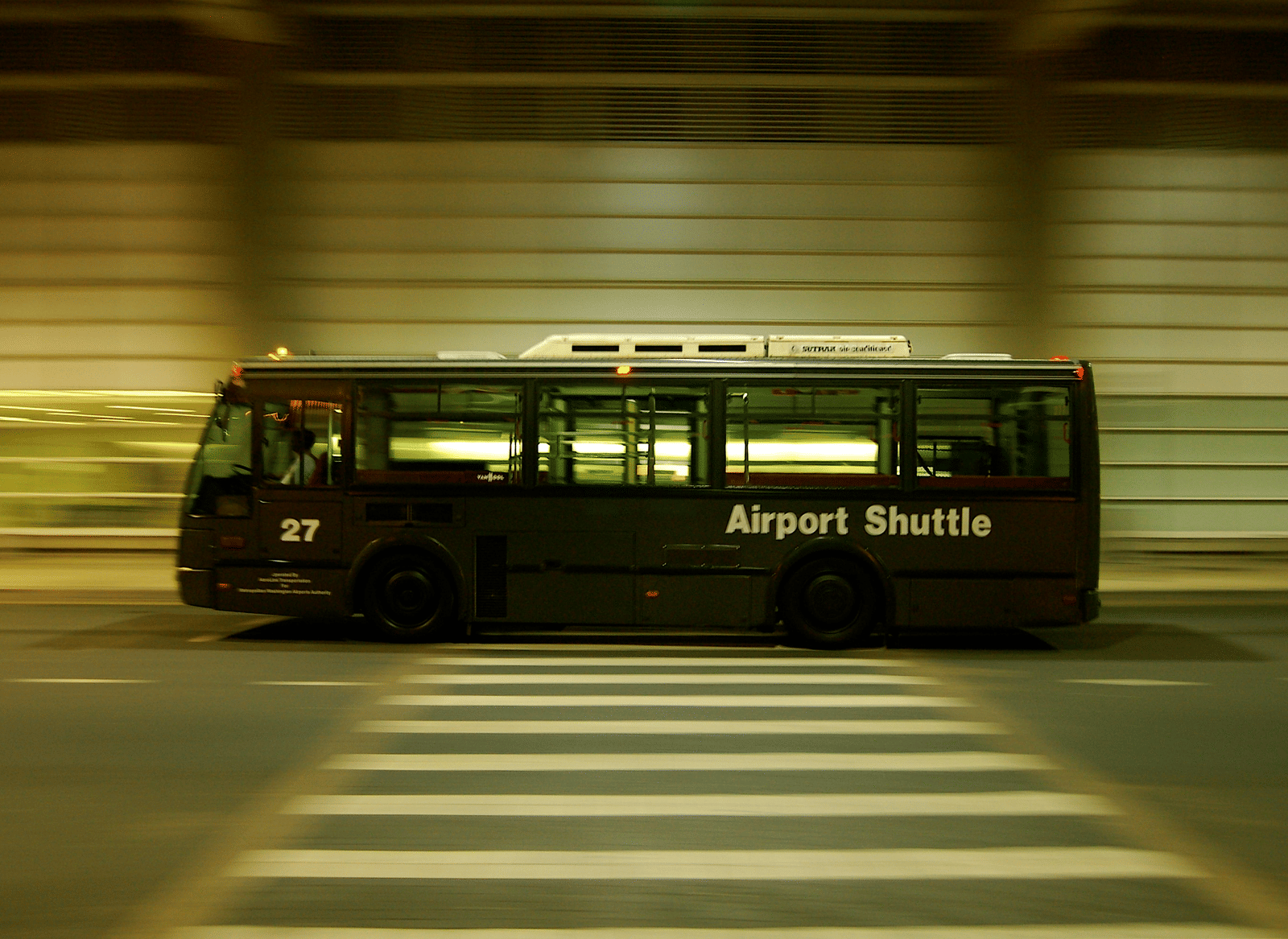 A fast-moving dark airport shuttle bus drives across a zebra crossing at night, with motion blur.