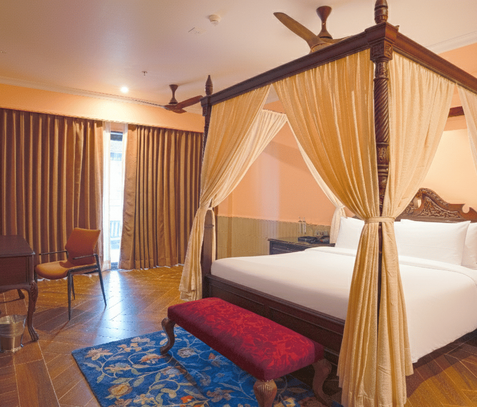 A bedroom with a 4-poster bed draped in sheer curtains, wooden flooring, cushioned bench, sliding curtains and a small dining table set near the window at MAYFAIR Bay Resort, Paradeep.