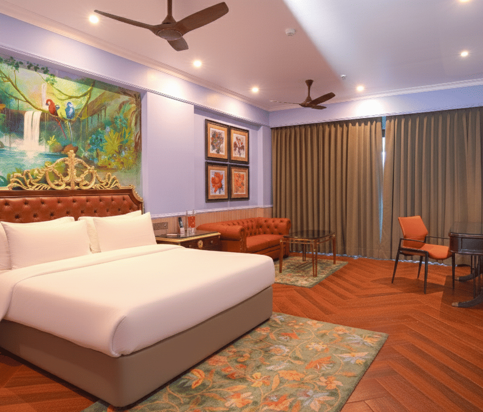 A well-lit room featuring a plush king size bed, artistic headboard, wall paintings, a work desk and a chair, wooden flooring and ceiling fan with floor-to-ceiling curtains at MAYFAIR Bay Resort, Paradeep.