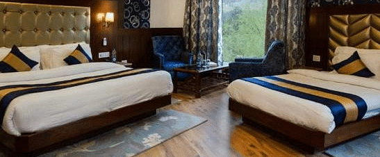 A spacious, well-lit hotel room at La Serene Valley by DLS Hotels in Manali, India. The room features two beds, a sitting area, and large windows offering natural light and views of the surrounding mountains | DLS La Serene Manali