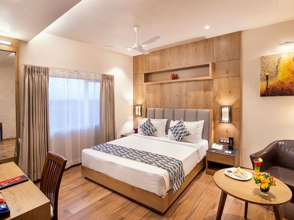 A hotel room showcasing a double bed with white and grey bedding, a wooden bedside table with a lamp, a work desk area with a chair and television, and a small round table with a brown armchair, complemented by wooden flooring at Lords Inn Motikhavdi