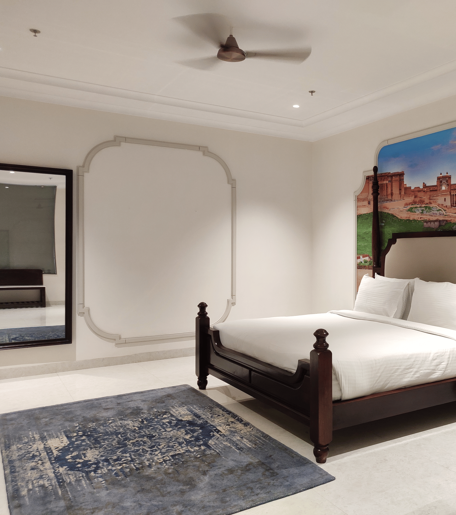 A wide shot of a bedroom at Anant Mahal, Jaipur featuring a king size bed, a blue patterned rug, a mirror, a ceiling fan, and a large mural behind the headboard.