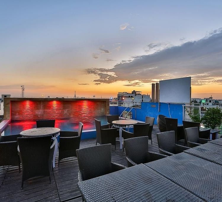 A rooftop terrace during sunset with wicker furniture, a lit pool and a view of the city skyline - TGI Apple Inn, Jaipur 