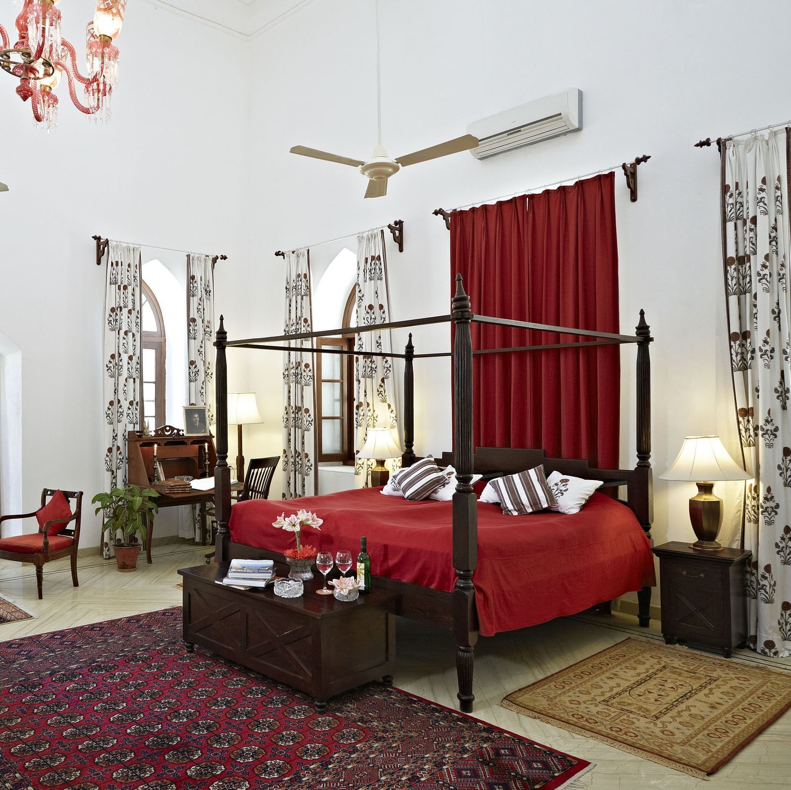A spacious room featuring a four-poster bed with linens, seating arrangements, decorative rugs, and arched windows with draped curtains- Shahpura Bagh.