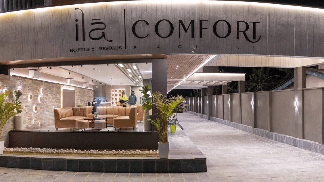 Hotel entrance at night with illuminated brand signage, a glass-fronted lobby lounge and a lamp-lit walled driveway at Ila Comfort, Baglung.