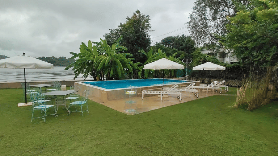 Outdoor pool area at Ram Pratap Palace, Udaipur, featuring sun loungers, umbrellas, a dining table with chairs, lush greenery, and a lake view in the background.