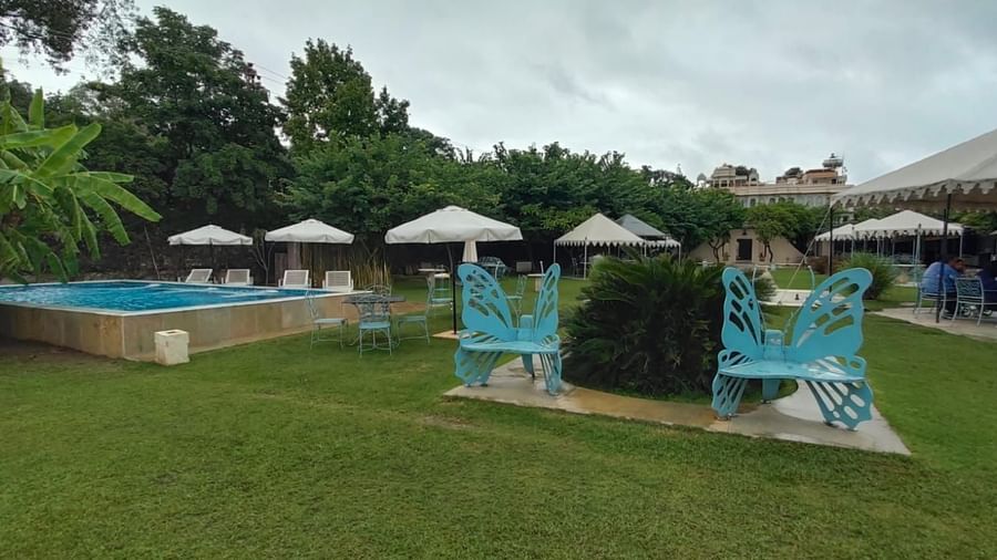 Garden area at Ram Pratap Palace, Udaipur featuring a swimming pool, butterfly-shaped bench, white umbrellas, and guests dining under canopy tents.