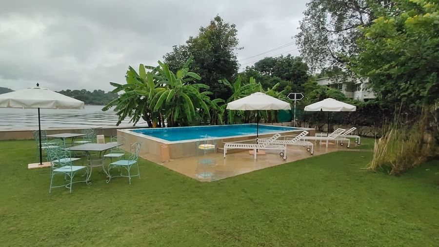 Outdoor pool area at Ram Pratap Palace, Udaipur, featuring sun loungers, umbrellas, a dining table with chairs, lush greenery, and a lake view in the background.