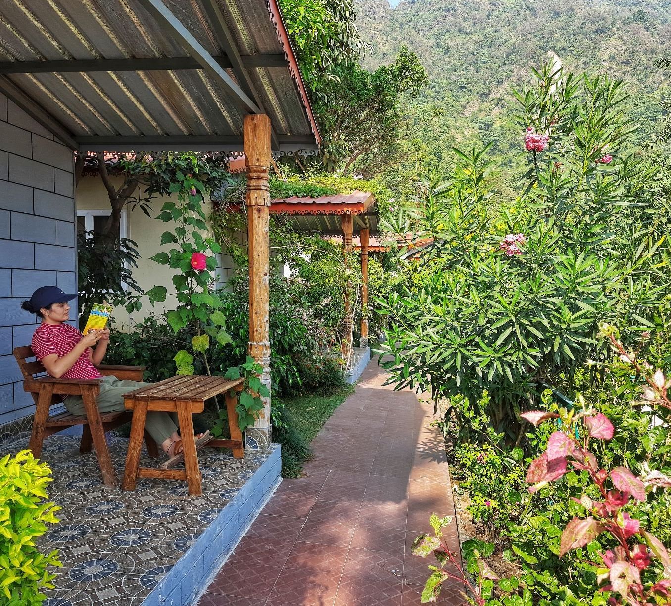 a person reading a book outside one of the room with a garden in front of them - Neer Ganga Resorts