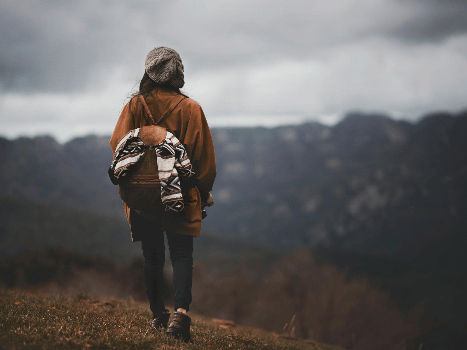 A lone hiker with a backpack walking along a mountain trail on a cloudy day, surrounded by misty hills and rugged natural scenery.
