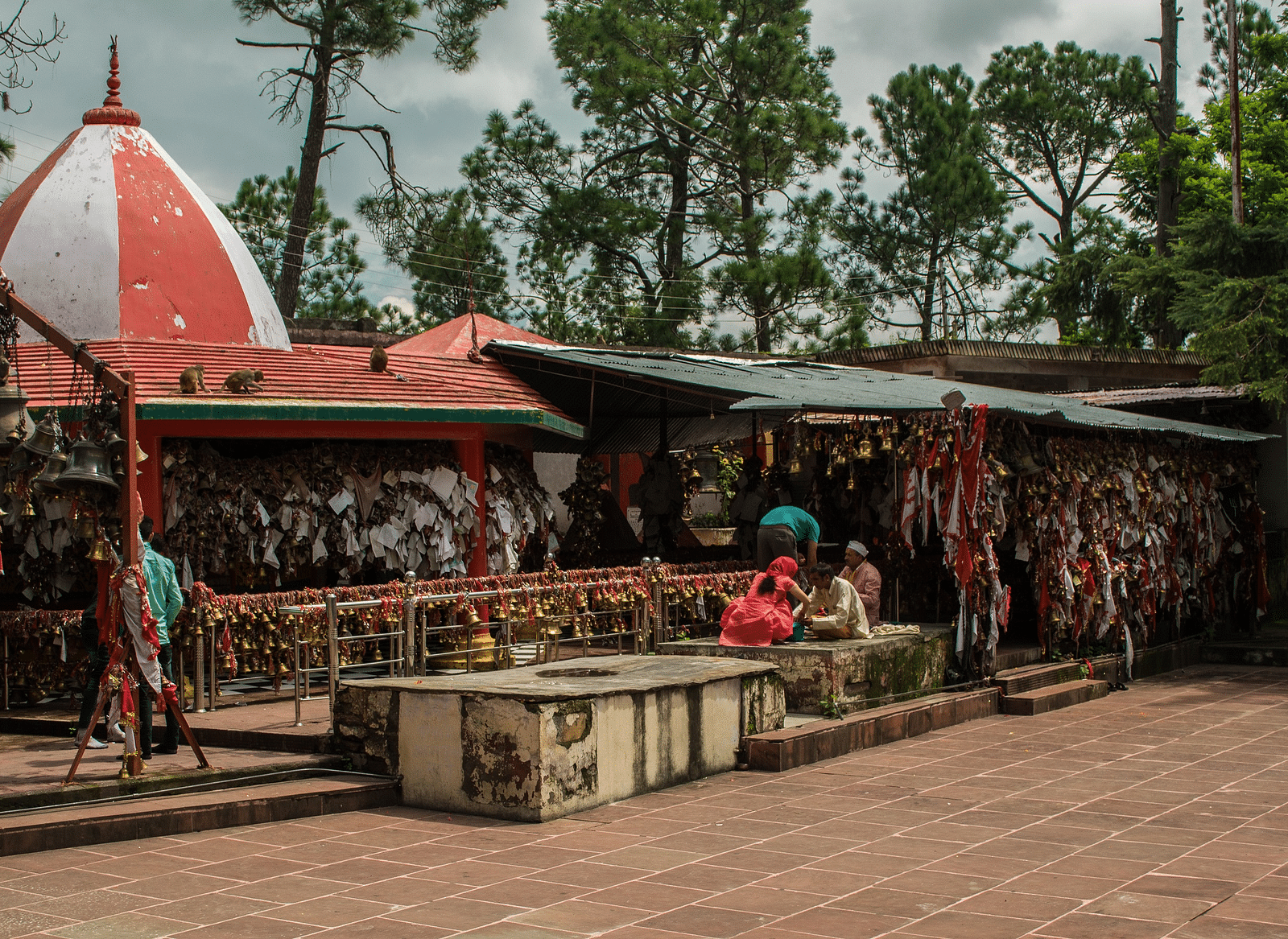 A colourful temple with hanging offerings, surrounded by trees under a cloudy sky.