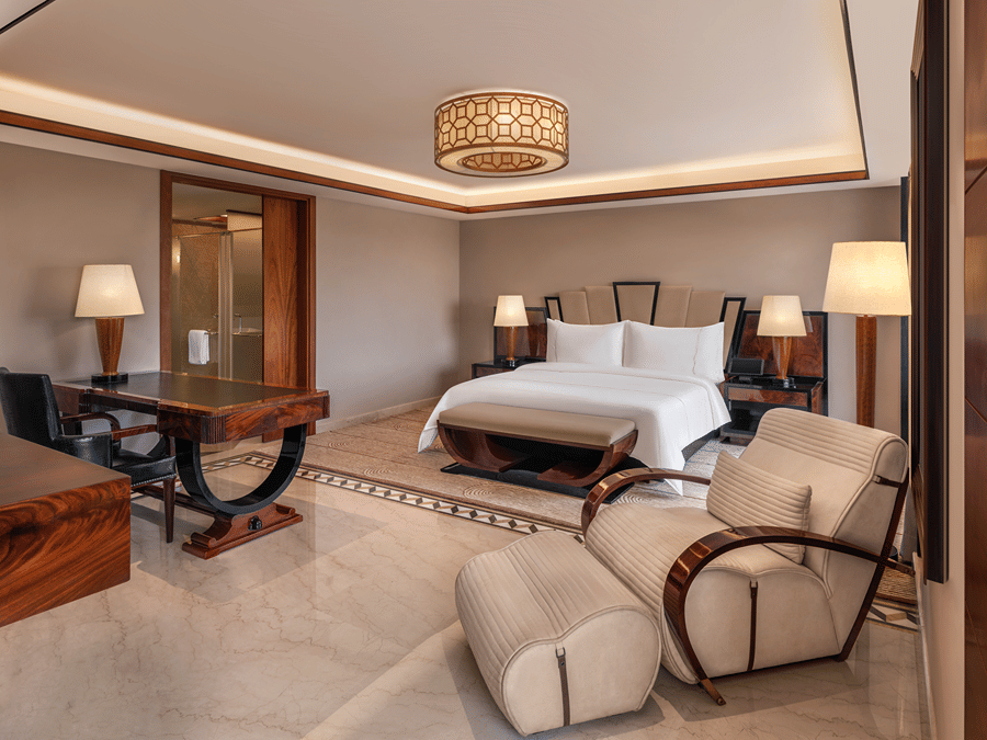 Presidential Suite bedroom with a king bed, art deco headboard, writing desk, cream lounge chairs and a marble floor, with an en-suite bathroom in the background.