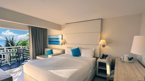A neatly set room at The Soco Hotel featuring a large white bed with blue pillows, a lit lamp, and a balcony view of the ocean.