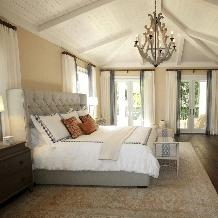 A spacious, elegant bedroom with a tufted bed, wooden furniture, chandelier, large windows, and soft natural light creating a warm, airy ambience.