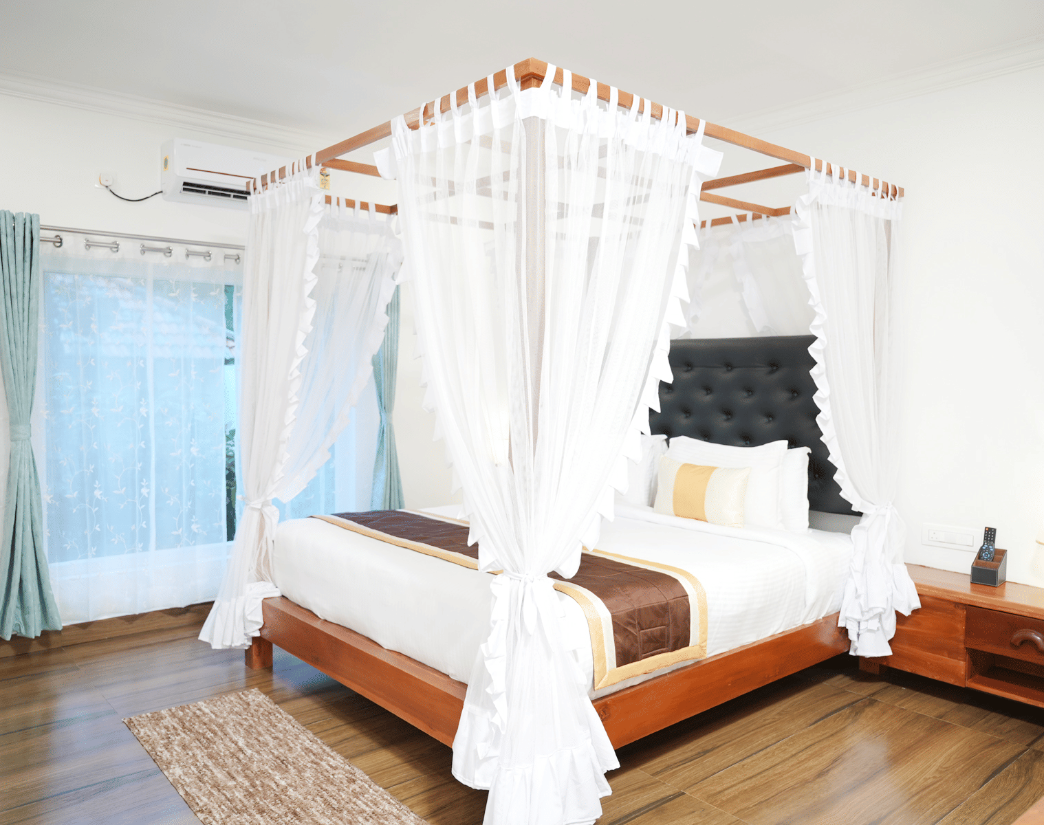 A luxurious hotel bedroom featuring a four-poster wooden bed with white drapery and a brightly lit window at Coorg Orange Blossom Resort & Spa.