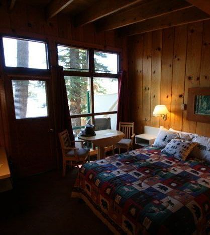 A rustic cabin room with wood paneling features a bed with a colorful quilt and a large window. - Kit Carson Lodge & Restaurant