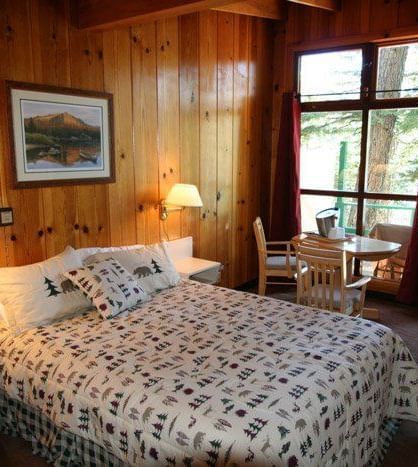 A room featuring a bed with patterned linens, a small table and chairs, and a view of the outdoors through a window. - Kit Carson Lodge