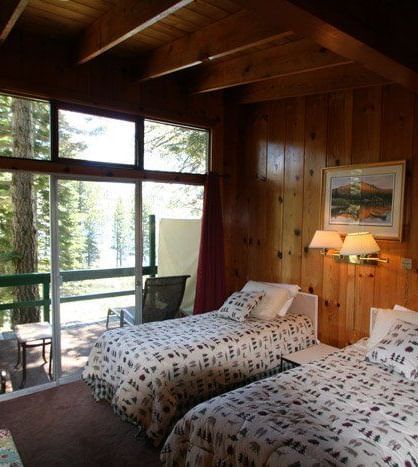 A rustic cabin room with two twin beds featuring patterned comforters, warm wood paneling and a large window. - Kit Carson Lodge & Restaurant 