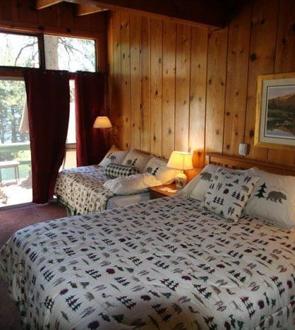 A cosy cabin room with two beds and patterned comforters, warm wood paneling, a framed picture and  glass door leading to a balcony. - Kit Carson Lodge & Restaurant