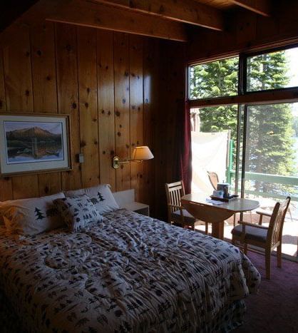 A bedroom with wood paneling, featuring a bed with a patterned comforter, two bedside lamps and a framed picture. -  Kit Carson Lodge & Restaurant