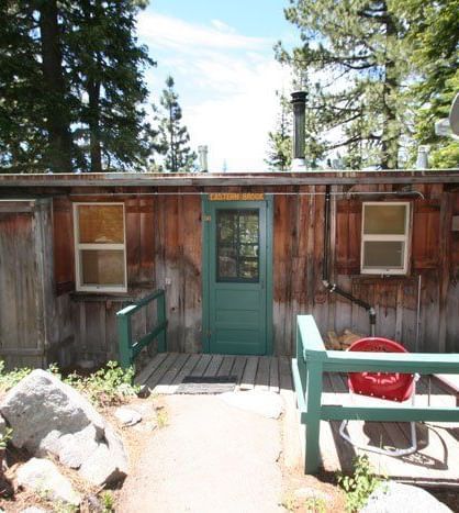 A charming wooden cabin with multiple windows and a small green porch, nestled in a sunny forest setting | Eastern Brook
