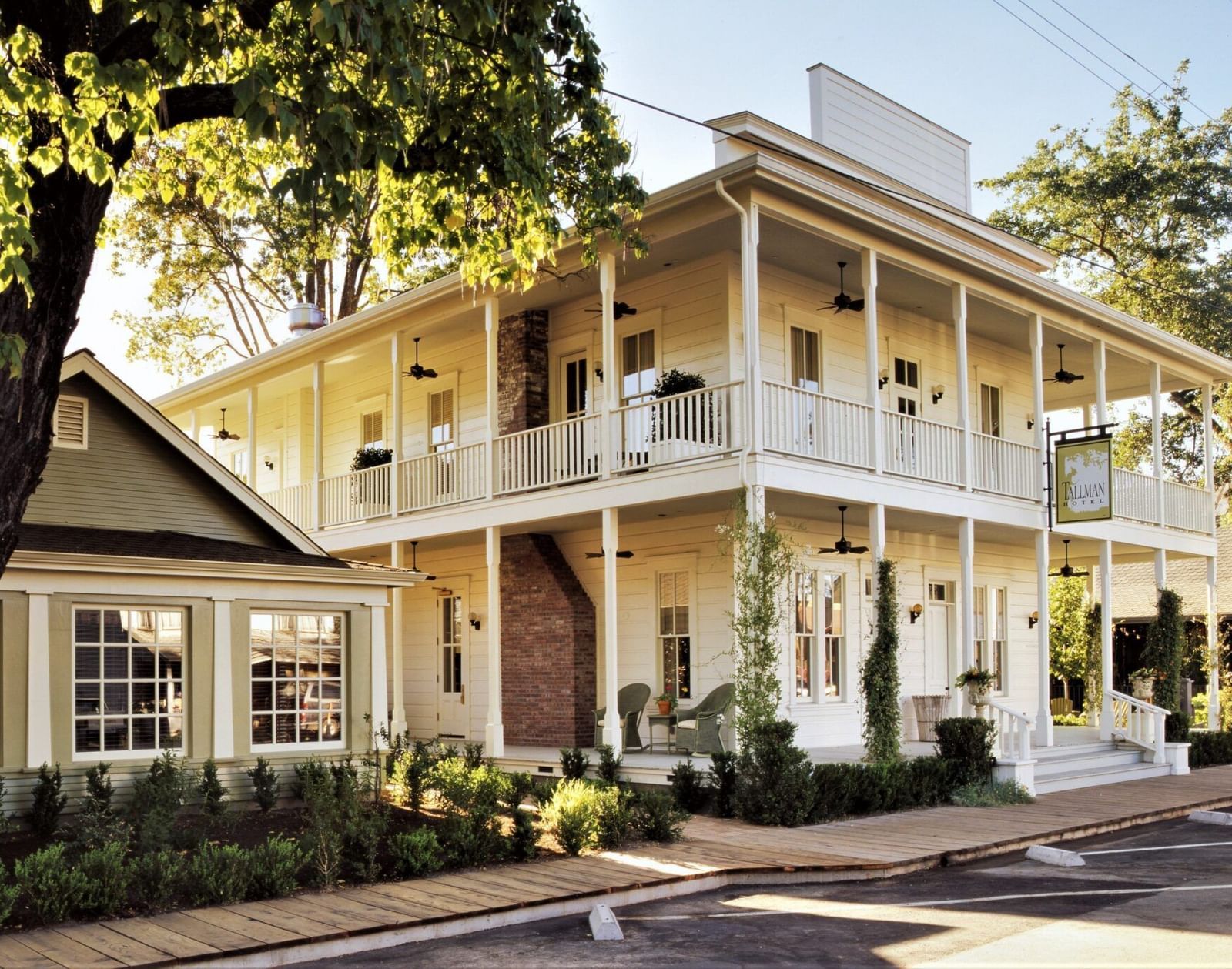 Corner view of Tallman Hotel showing two-story white exterior with wraparound balconies and surrounding greenery.