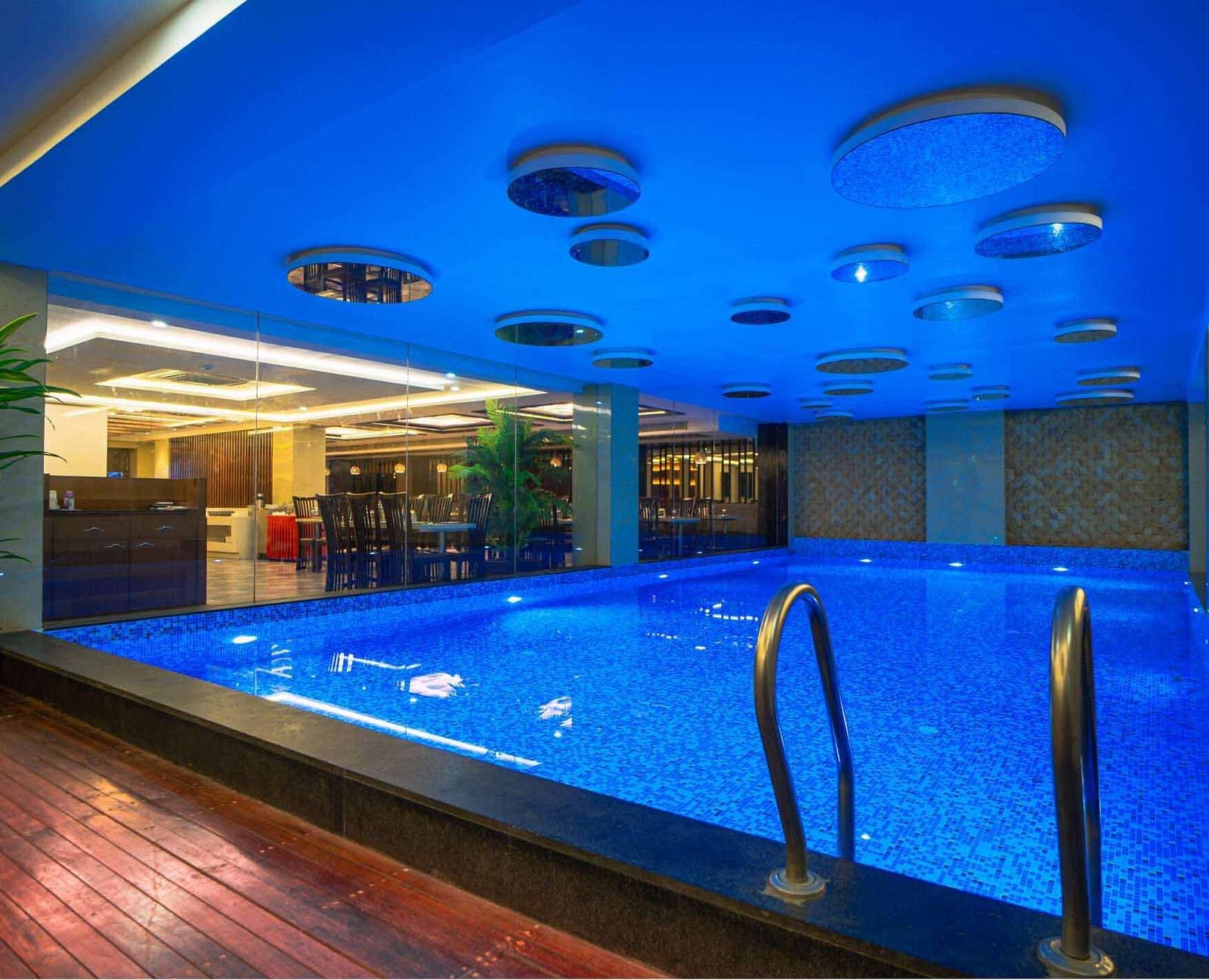 An indoor swimming pool with blue water and a blue ceiling, illuminated by warm lighting at Mansha Hotel Regency.