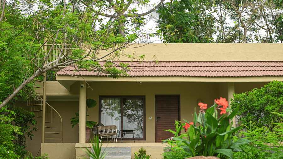 Exterior view of a cottage at Fireflies Resort Kabini surrounded by lush greenery and tropical plants in a serene natural setting.