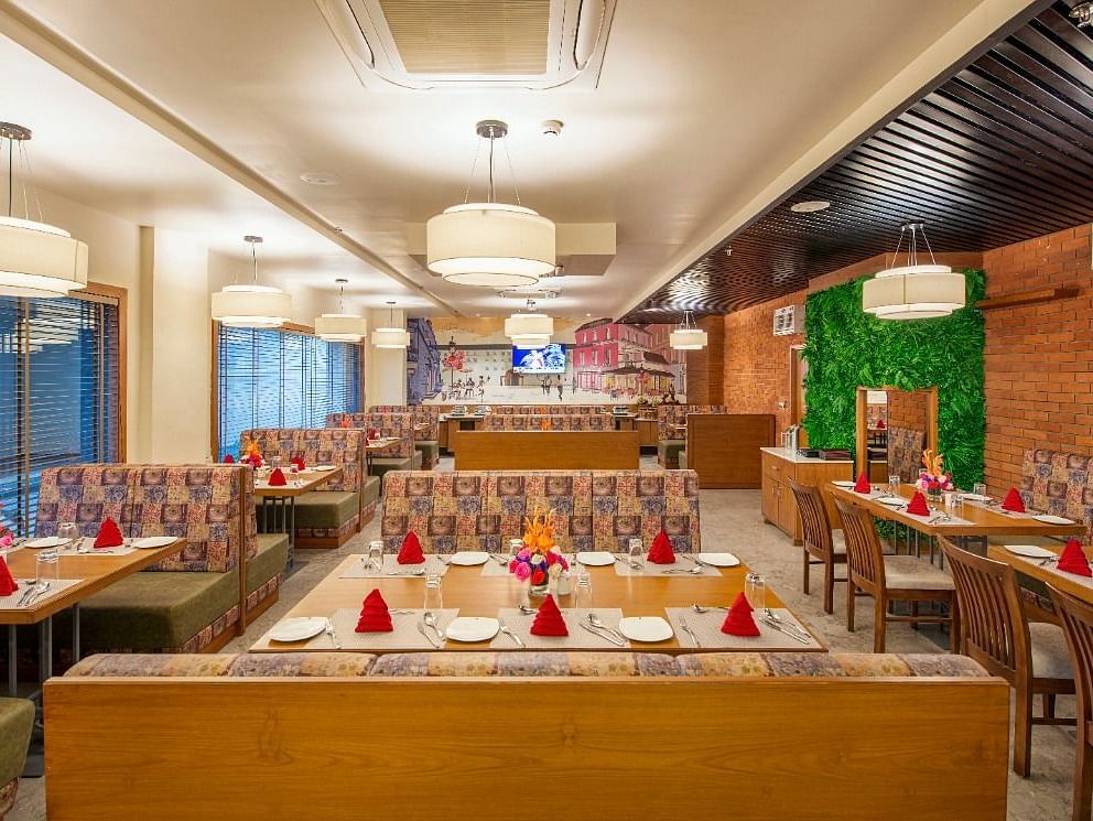 A warmly lit and inviting restaurant dining area with numerous tables set for meals, featuring patterned booth seating and wooden chairs, red napkins, and floral centrepieces, under soft pendant lighting - Lords Inn Motikhavdi, Jamnagar.