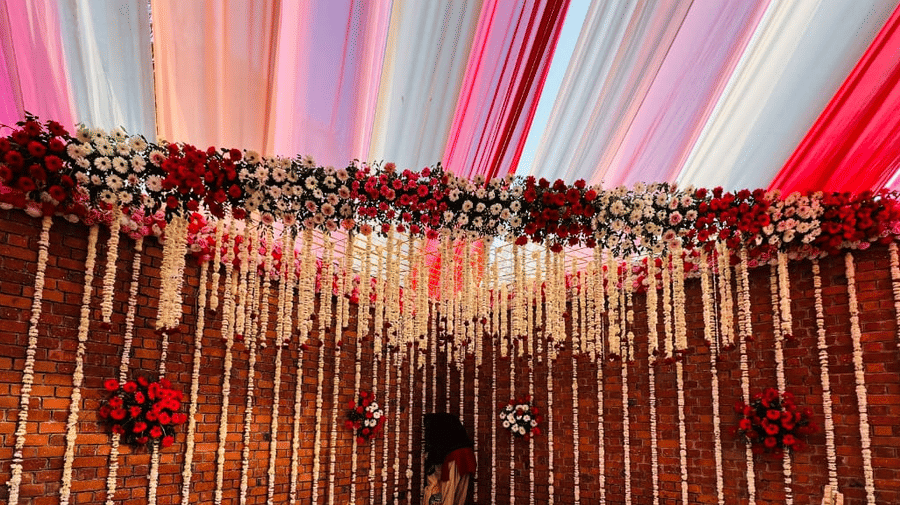 A covered outdoor event space with a stage backdrop made of thin wooden slats, draped with flowing pink and sheer white fabric and dark red flowers | Nandan Resort
