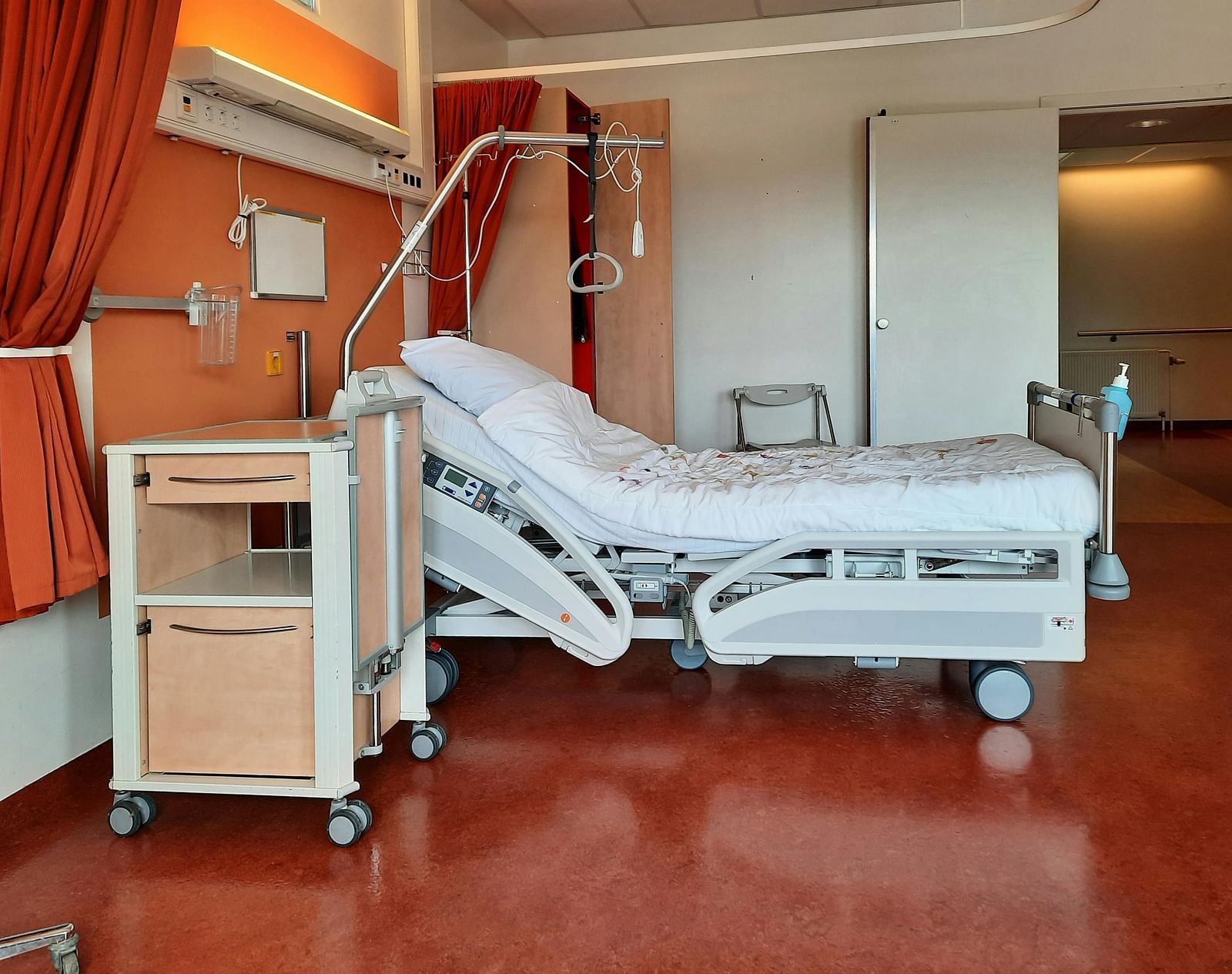 A hospital room with a white bed, bedside table, and medical equipment on a red floor.