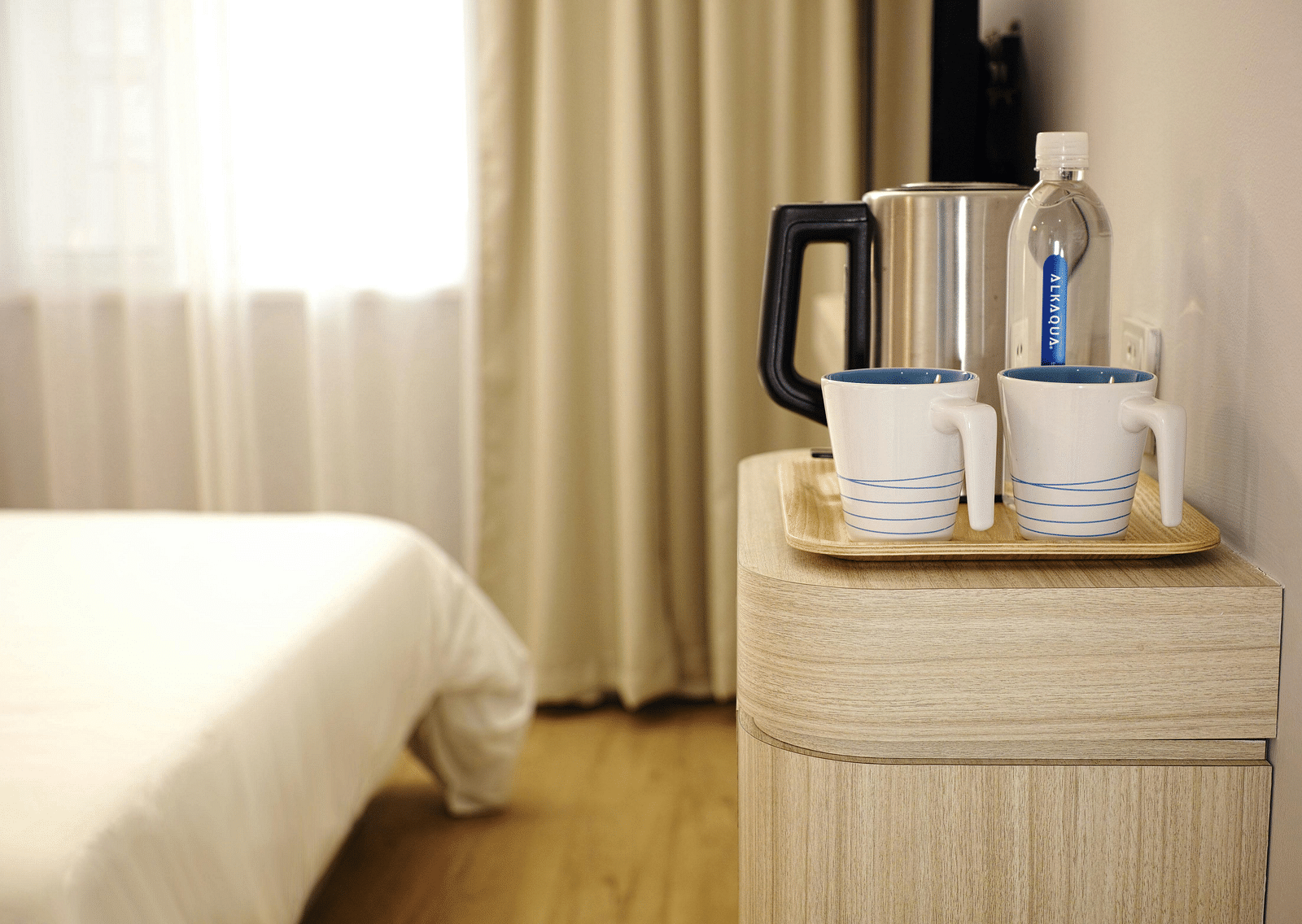 Two white ceramic mugs on brown wooden tray placed on a table next to a hotel bed