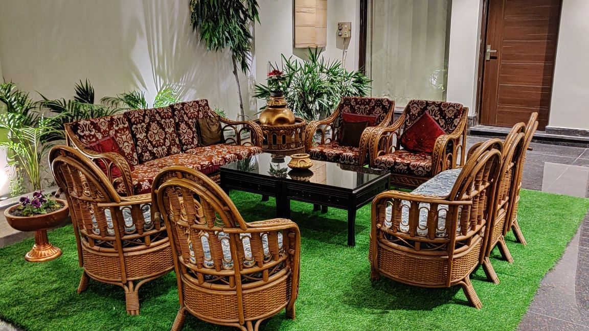 An interior shot of a lounge or seating area at Hotel Exotic Amritsar, featuring a set of wicker furniture including a sofa, chairs, and a table, all arranged on a green rug. 