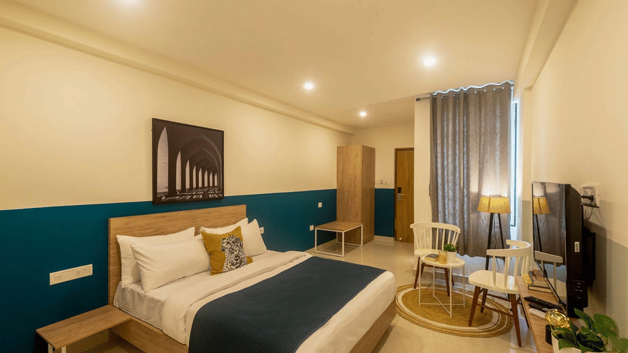 A wide-angle view of a hotel room showing a queen-sized bed, a wooden headboard, a small round table with two chairs, and a large window.