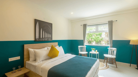 A bright hotel room with a queen bed, minimalist wooden furniture, and a pair of white chairs near a window, with a large framed art piece above the bed.