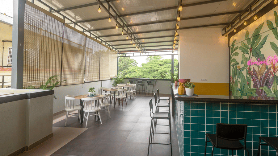 A perspective view of a long, narrow outdoor balcony or cafe area with white tables and chairs, looking toward the teal tiled bar and mural.