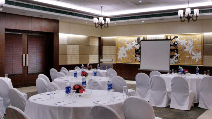 Banquet hall setup at Goldfinch Bengaluru featuring round dining tables with white tablecloths, chairs, and a screen.