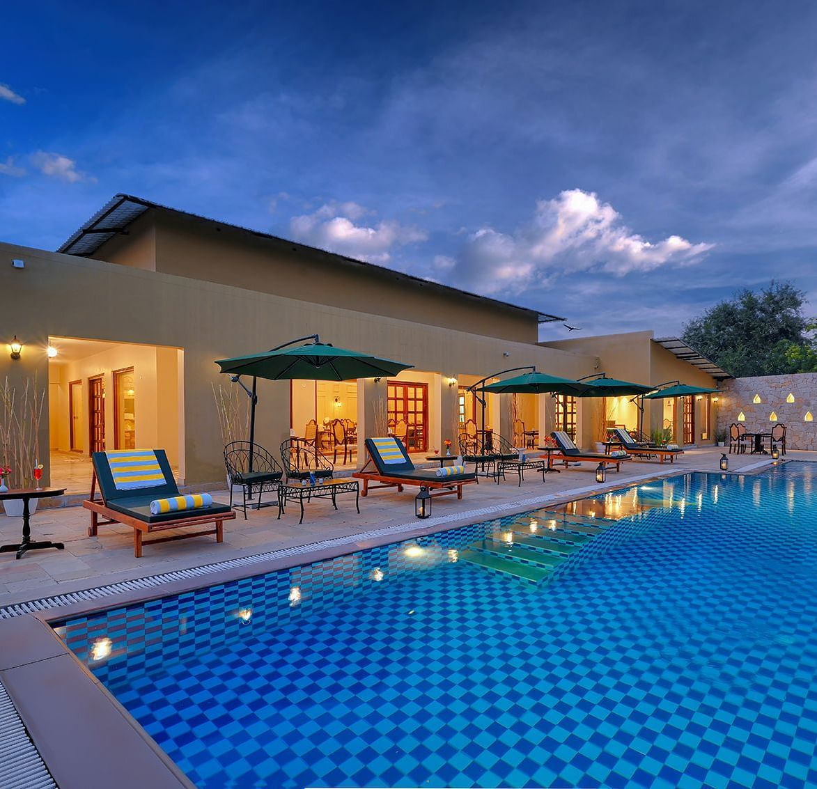 Exterior view of a resort pool with umbrellas and lounge chairs under a cloudy evening sky - Singhvilas, Ranthambore