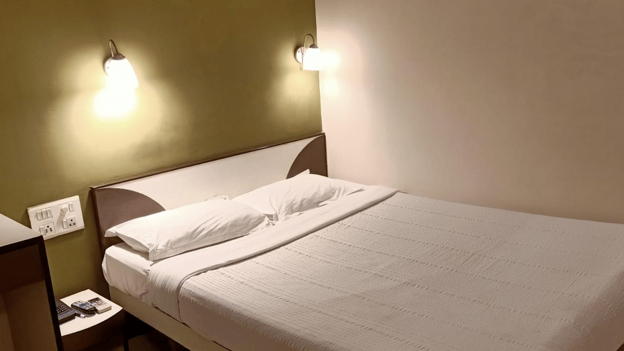 A Standard AC Room at Hotel Pandian, featuring a neatly-made bed with white sheets, 2 pillows, 2 wall lamps, and a nightstand.