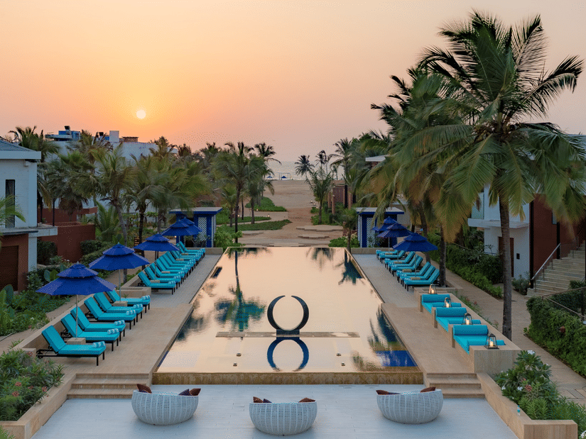 an aerial view of the swimming pool at Azaya Beach Resort, the best stay in South Goa, with loungers and coconut trees lined next to it and the sun setting in the background.