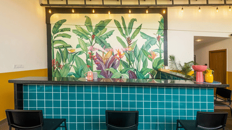 A straight-on shot of a bar counter covered in teal square tiles, backed by a colorful mural of oversized tropical plants and pink flowers.