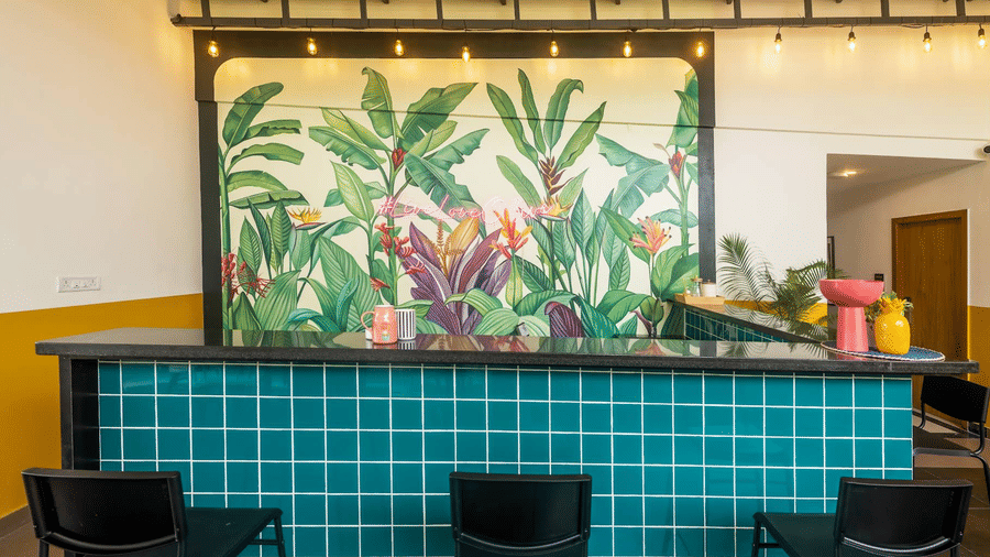 A straight-on shot of a bar counter covered in teal square tiles, backed by a colorful mural of oversized tropical plants and pink flowers.
