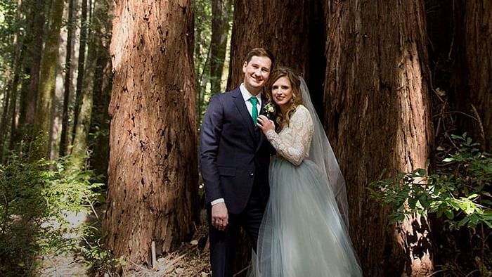 Bride and groom standing beneath towering trees, bathed in soft light