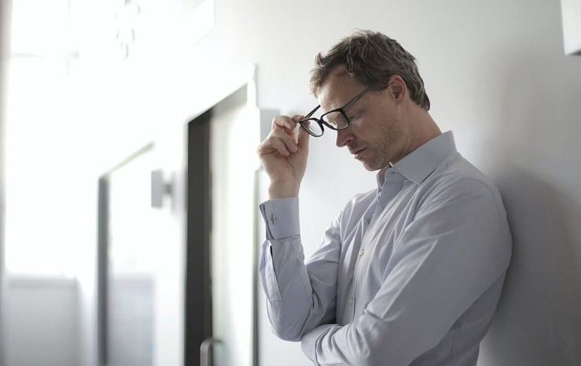 A man in a light blue shirt leans against a white wall, holding his glasses, appearing stressed or deep in thought at YO1 Longevity and Health Resorts