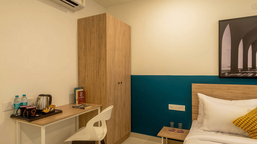 A corner view of the hotel room showing a wooden wardrobe, a small desk with a chair, and a coffee-making station with bottled water.