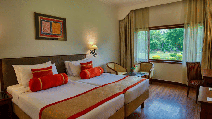 The Deluxe Twin Garden View room at Clarks Khajuraho featuring twin beds pushed together with crisp linens and decorative bolsters along with a wooden work desk, a large window overlooking a lush green garden, and warm accent lighting from bedside lamps.