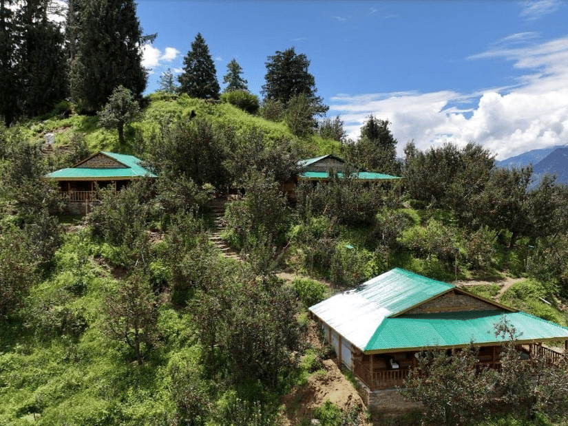 A beautiful landscape featuring 3 cottages amid lush green vegetation at Amara Upepo - The Sky Village, Manali, with a clear blue sky in the background.
